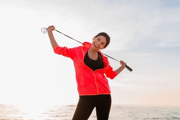 Junge attraktive schlanke Frau, die Sportübungen im Morgensonnenaufgang am Meeresstrand in der Sportkleidung, im gesunden Lebensstil, trägt rosa Windbreakerjacke tut, Springseil hält