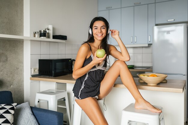 Junge attraktive sexy Frau, die Frühstück in der Küche am Morgen, Apfel aß, lächelnd, glücklich, positiv, gesunder Lebensstil, Musik auf Kopfhörern hörend