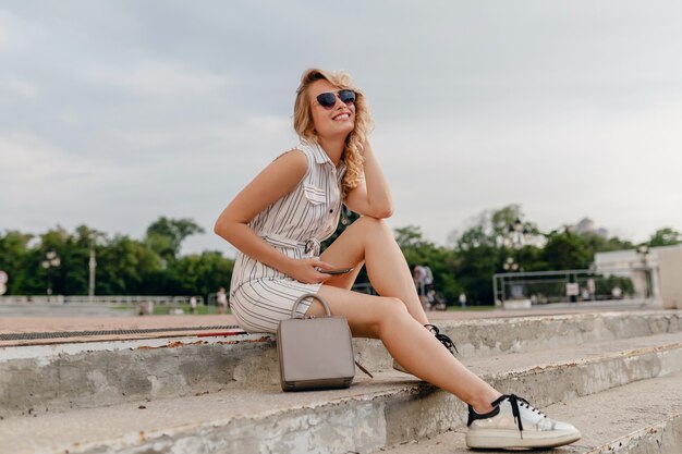 Junge attraktive stilvolle blonde Frau, die in der Stadtstraße im Sommermodeartkleid sitzt, das Sonnenbrille, Geldbörse, silberne Turnschuhe trägt