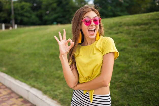 Junge attraktive stilvolle lächelnde Frau, die Spaß im Stadtpark hat, positiv, emotional, tragendes gelbes Oberteil, gestreiften Minirock, rosa Sonnenbrille, Sommerart-Modetrend, zeigt gutes Zeichen, bunt