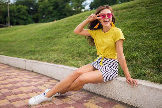 Junge attraktive stilvolle lächelnde Frau, die Spaß im Stadtpark hat, positiv, emotional, tragendes gelbes Oberteil, gestreiften Minirock, rosa Sonnenbrille, weiße Turnschuhe, Sommerart-Modetrend, Friedenszeichen