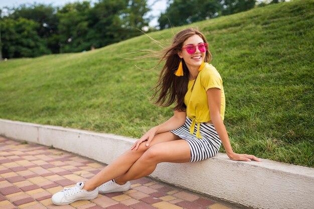 Junge attraktive stilvolle lächelnde Frau, die Spaß im Stadtpark hat, positiv, emotional, tragendes gelbes Oberteil, gestreiften Minirock, rosa Sonnenbrille, weiße Turnschuhe, Sommerart-Modetrend, lange Beine