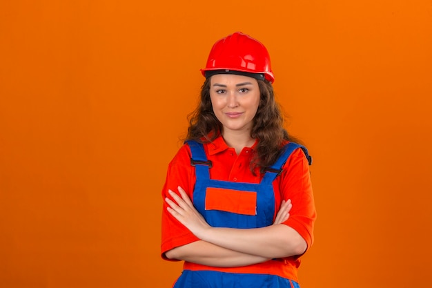 Junge Baumeisterfrau in Bauuniform und Sicherheitshelm stehend mit verschränkten Armen mit Lächeln auf Gesicht über isolierter orange Wand