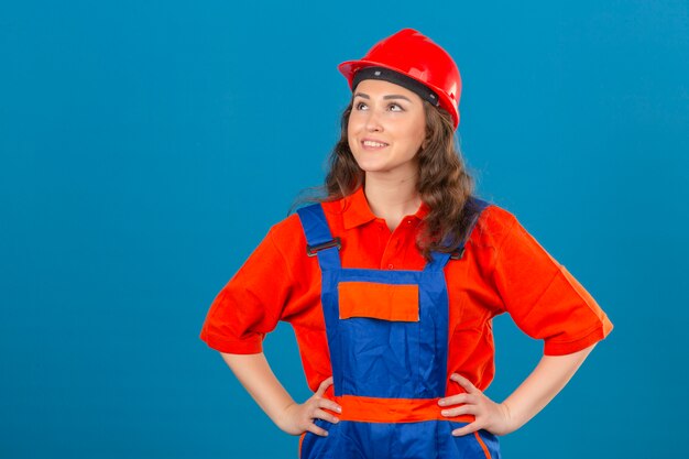 Junge Baumeisterfrau in der Bauuniform und im Sicherheitshelm, die lächelndes glückliches Gesicht über isolierter blauer Wand suchen