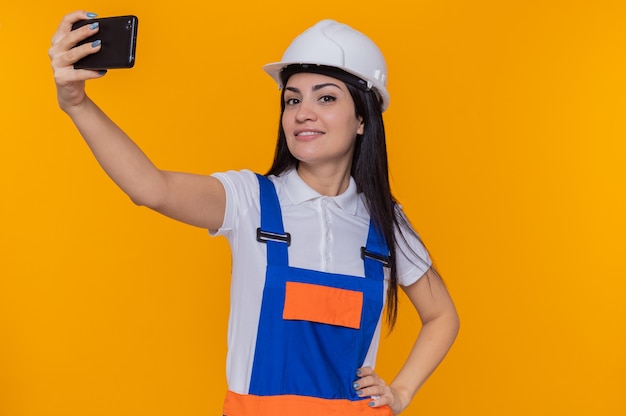 Junge Baumeisterfrau in der Bauuniform und im Sicherheitshelm, die selfie unter Verwendung des lächelnden selbstbewussten Smartphones des Smartphones über der orangefarbenen Wand tun