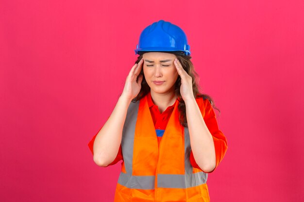 Junge Baumeisterfrau in der Bauuniform und im Sicherheitshelm, die unwohl suchen, ihren Kopf berührend, der starke Kopfschmerzen über isolierter rosa Wand hat