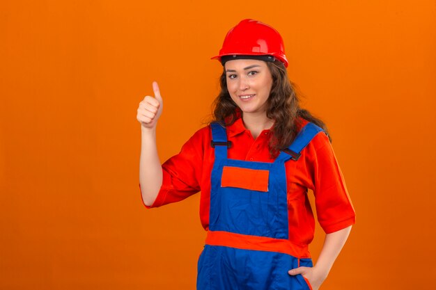 Junge Baumeisterfrau in der Bauuniform und im Sicherheitshelm lächelnd, die Daumen oben über isolierter orange Wand zeigen