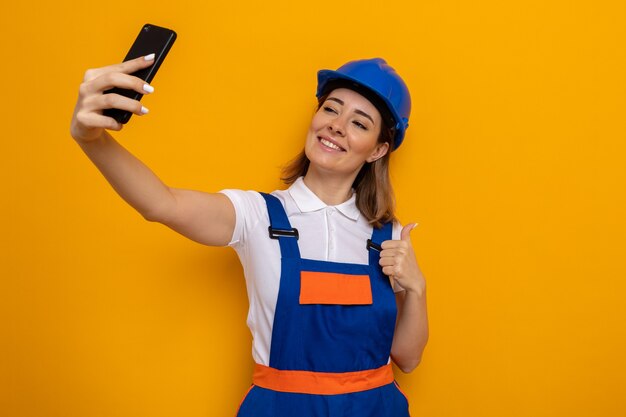 Junge Baumeisterin in Bauuniform und Schutzhelm, die fröhlich glücklich und positiv lächelt und Selfie mit dem Smartphone macht, das Daumen nach oben über oranger Wand zeigt