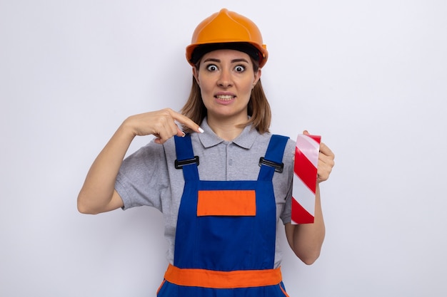 Junge Baumeisterin in Bauuniform und Schutzhelm, die Klebeband hält und mit dem Zeigefinger darauf zeigt, dass sie verwirrt über der weißen Wand steht?