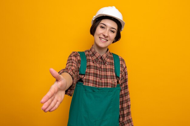 Junge Baumeisterin in Bauuniform und Schutzhelm, die lächelnd freundlich aussieht und die Hand anbietet, die Grußgeste macht
