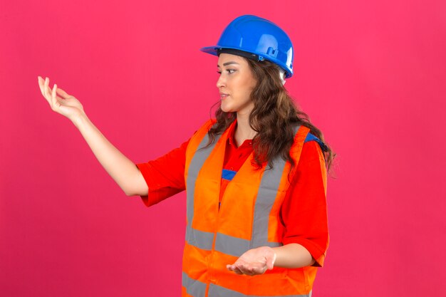 Junge Baumeisterin in Bauuniform und Sicherheitshelm gestikulierte enttäuscht stehend mit den Händen, die über isolierte rosa Wand erhoben wurden