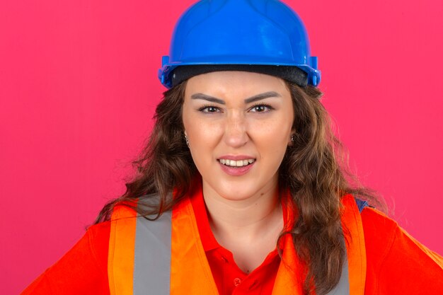 Junge Baumeisterin in Bauuniform und Sicherheitshelm mit wütendem Ausdruck über isolierter rosa Wand