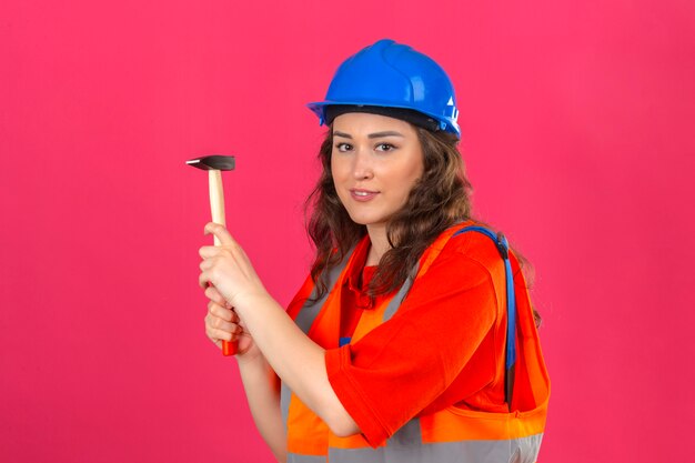 Junge Baumeisterin in Bauuniform und Sicherheitshelm stehend mit Hammer, der mit Lächeln an der Kamera über isolierter rosa Wand schaut