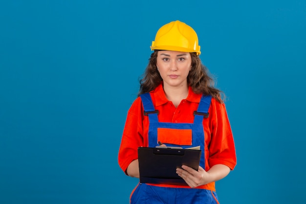 Junge Baumeisterin in Bauuniform und Sicherheitshelm stehend mit Klemmbrett in Händen mit ernstem Gesicht über isolierter blauer Wand