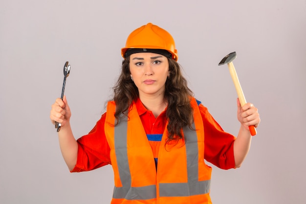 Junge Baumeisterin in Bauuniform und Sicherheitshelm stehend mit Schraubenschlüssel und Hammer mit ernstem Gesicht über isolierter weißer Wand