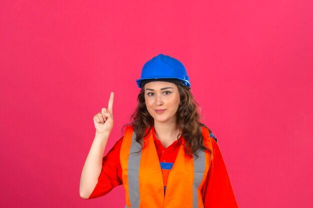 Junge Baumeisterin in der Bauuniform und im Sicherheitshelm, der mit neuem Ideenkonzept des Fingers über isolierte rosa Wand zeigt