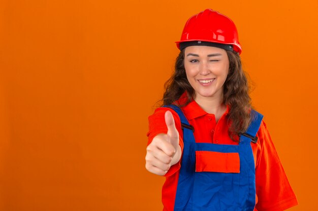 Junge Baumeisterin in der Bauuniform und im Sicherheitshelm, die Daumen hoch lächelnd fröhlich über isolierte orange Wand zwinkert