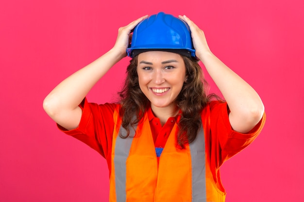 Junge Baumeisterin in der Bauuniform und im Sicherheitshelm, die überraschten und glücklichen berührenden Helm mit den Händen über der isolierten rosa Wand suchen