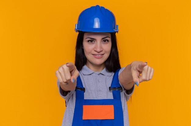 Junge Baumeisterin in der Bauuniform und im Sicherheitshelm lächelnd, die sicher zeigen, dass mit beiden Händen Zeigefinger vorne über orange Wand stehen