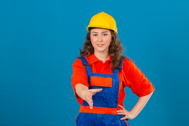 Junge Baumeisterin in der Bauuniform und im Sicherheitshelm mit dem Lächeln, das die Hand anbietet, die Grußgeste über isolierte blaue Wand macht