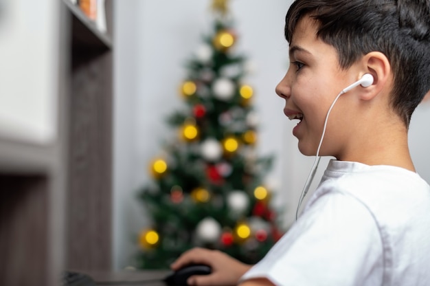 Junge benutzt PC mit Kopfhörern. Weihnachtsbaum an der Wand. Glückliches und aufgeregtes Gesicht