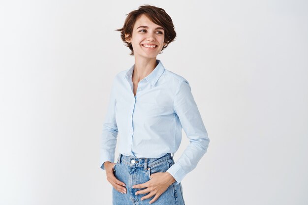 Junge Berufsfrau im blauen Kragenhemd, die hoffnungsvoll und glücklich beiseite schaut, auf den leeren Raum starrt und lächelt, auf weißer Wand stehend