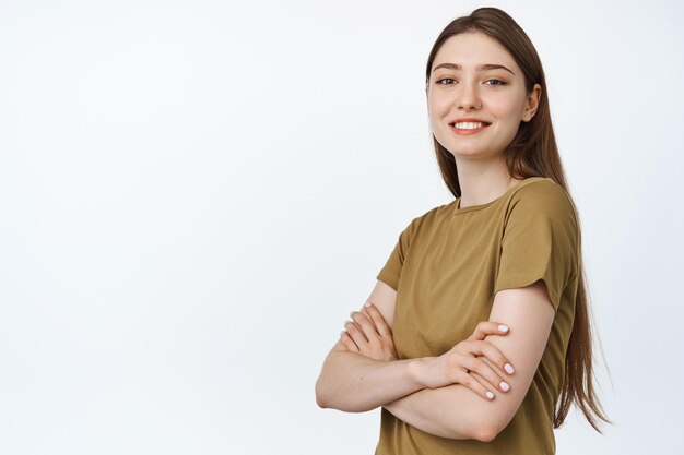 Junge Berufstätige. Lächelndes Mädchen verschränkt die Arme auf der Brust und schaut selbstbewusst in die Kamera, steht im T-Shirt vor weißem Hintergrund.