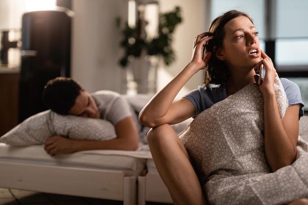 Kostenloses Foto junge besorgte frau, die an etwas denkt, während sie im schlafzimmer sitzt ihr freund schläft auf dem bett