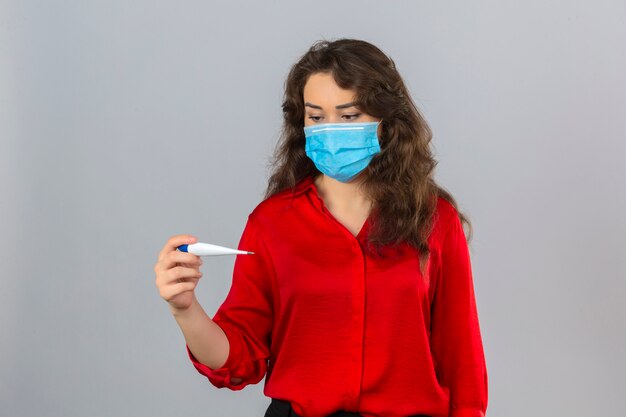 Junge besorgte Frau, die rote Bluse in der medizinischen Schutzmaske trägt, die digitales Thermometer in ihrer Hand über lokalisiertem weißem Hintergrund betrachtet