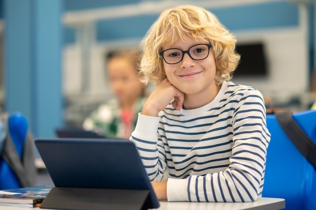 Junge blickt in die Kamera und sitzt mit Tablet am Schreibtisch