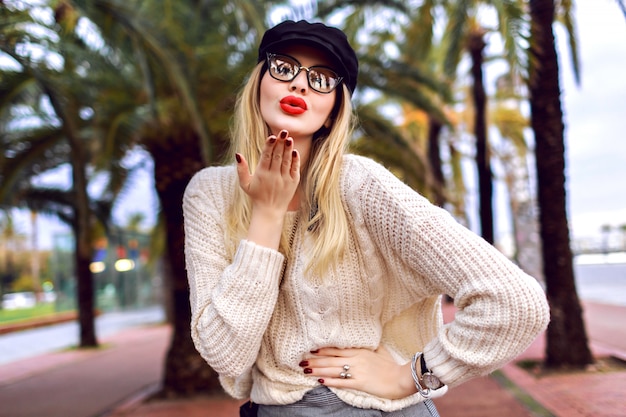 Junge blonde elegante stilvolle Frau, die Kuss sendet und auf Barcelona Straße mit Palmen aufwirft, kuscheligen Pullover, Mütze und klare Brille tragend, Modestil, Reisestimmung, Frühlingszeit.