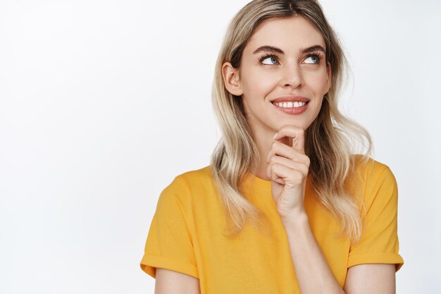 Junge blonde Frau, die lächelnd denkt und mit zufriedenem Gesicht zur Seite schaut, hat eine Idee, die in gelbem T-Shirt vor weißem Hintergrund steht