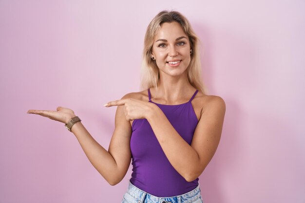 Junge blonde Frau, die über rosafarbenem Hintergrund steht und erstaunt in die Kamera lächelt, während sie mit der Hand präsentiert und mit dem Finger zeigt.