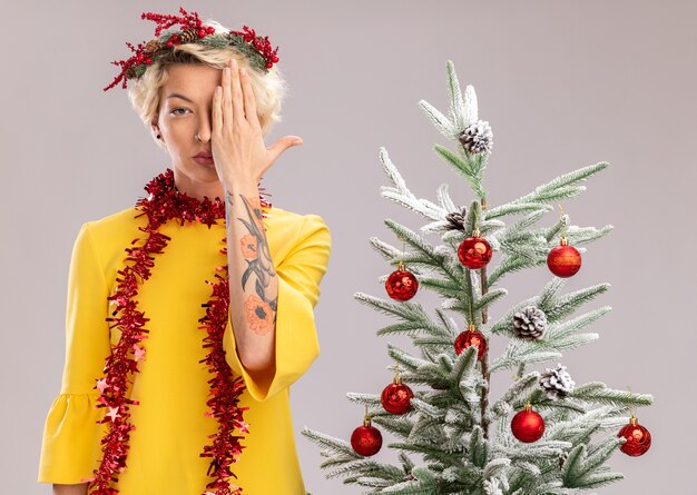 Junge blonde Frau, die Weihnachtskopfkranz und Lametta-Girlande um den Hals trägt, der nahe bei dekoriertem Weihnachtsbaum steht und Kamera betrachtet, die Hälfte des Gesichts mit Hand lokalisiert auf weißem Hintergrund bedeckt