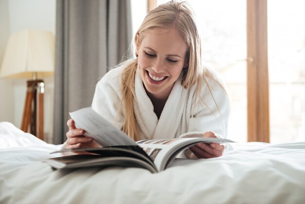 Junge blonde Frau, die Zeitschrift liest, während auf Bett liegend