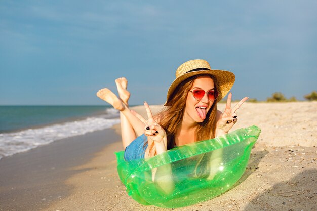 Junge blonde Frau entspannen und genießen Sie ihre Sommerferien, legen Sie sich auf Luftmatratze und nehmen Sie ein Sonnenbad, helle stilvolle Strandkleidung Hut und Sonnenbrille