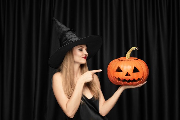 Junge blonde Frau im schwarzen Hut und im Kostüm auf schwarzem Hintergrund. Attraktives, sinnliches weibliches Model. Halloween, schwarzer Freitag, Cyber Montag, Verkauf, Herbst