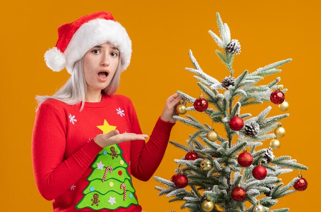 Junge blonde Frau im Weihnachtspullover und in der Weihnachtsmannmütze, die neben einem Weihnachtsbaum stehen, verwirrt, der sie mit Arm der Hand darstellt, die über orange Hintergrund ist