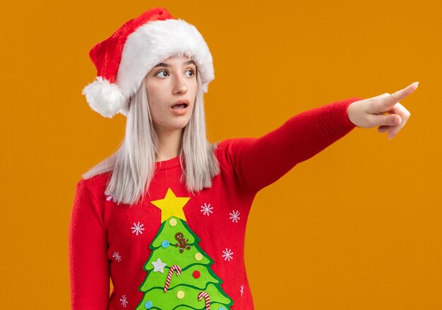 Junge blonde Frau im Weihnachtspullover und in der Weihnachtsmannmütze, die überrascht mit Zeigefinger auf etwas zeigen, das über orange Hintergrund steht