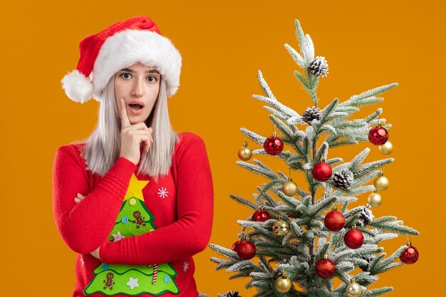 Junge blonde Frau im Weihnachtspullover und in der Weihnachtsmütze überrascht und verwirrt