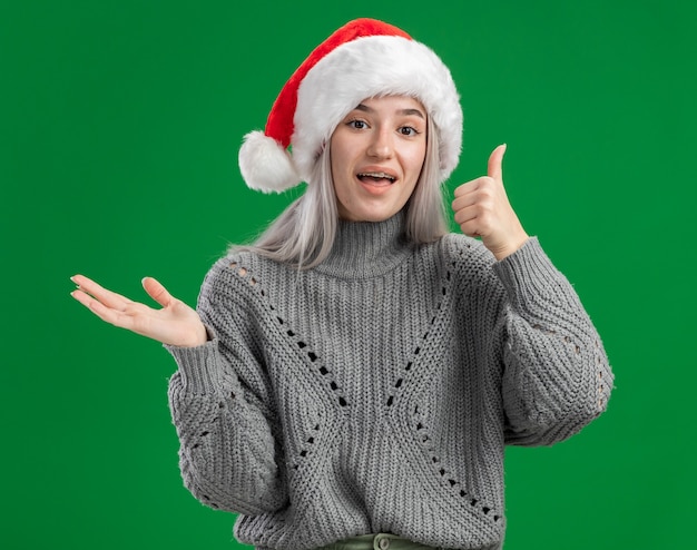 Junge blonde Frau im Winterpullover und in der Weihnachtsmannmütze, die Kamera glücklich und fröhlich zeigt, zeigt Daumen hoch, die Kopienraum mit Arm der Hand stehen über grünem Hintergrund darstellen