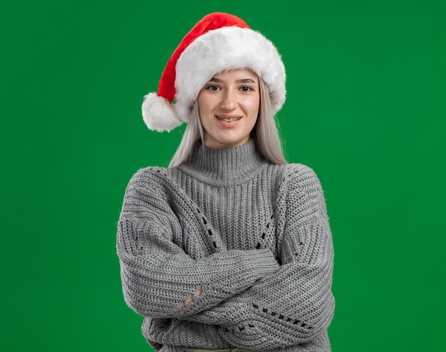 Junge blonde Frau im Winterpullover und in der Weihnachtsmannmütze, die Kamera mit Lächeln auf Gesicht mit verschränkten Armen betrachten, die über grünem Hintergrund stehen