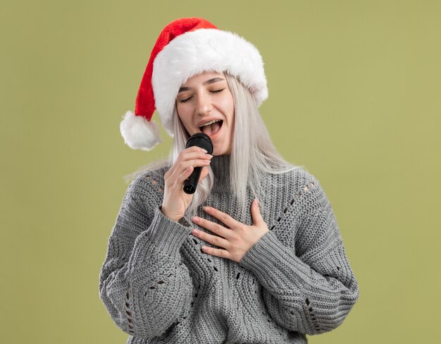 Junge blonde Frau im Winterpullover und in der Weihnachtsmannmütze, die Mikrofon hält, das mit geschlossenen Augen glücklich und positiv singt
