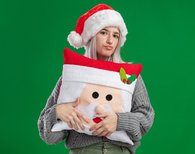 Junge blonde Frau im Winterpullover und in der Weihnachtsmannmütze, die Weihnachtskissen halten, das Kamera mit stirnrunzelndem Gesicht betrachtet, das über grünem Hintergrund nicht steht