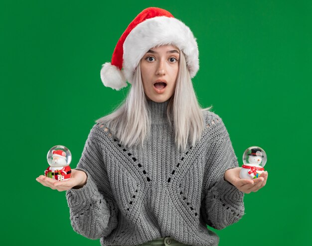 Kostenloses Foto junge blonde frau im winterpullover und in der weihnachtsmannmütze, die weihnachtsspielzeug-schneekugeln halten, die kamera betrachten, die über grünem hintergrund erstaunt und überrascht steht