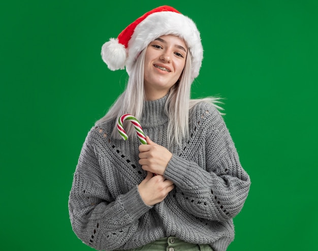 junge blonde Frau im Winterpullover und in der Weihnachtsmannmütze, die Zuckerstange hält, die Kamera glücklich und positiv lächelnd steht über grünem Hintergrund