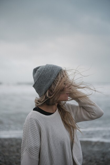 Junge blonde Frau in der Depression mit einem Hut, der auf dem Strand in einem windigen Wetter geht