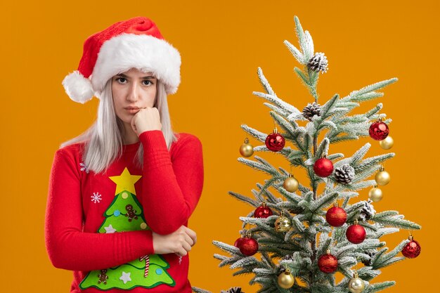 Junge blonde Frau in Weihnachtspullover und Weihnachtsmütze mit nachdenklichem Ausdruck denken