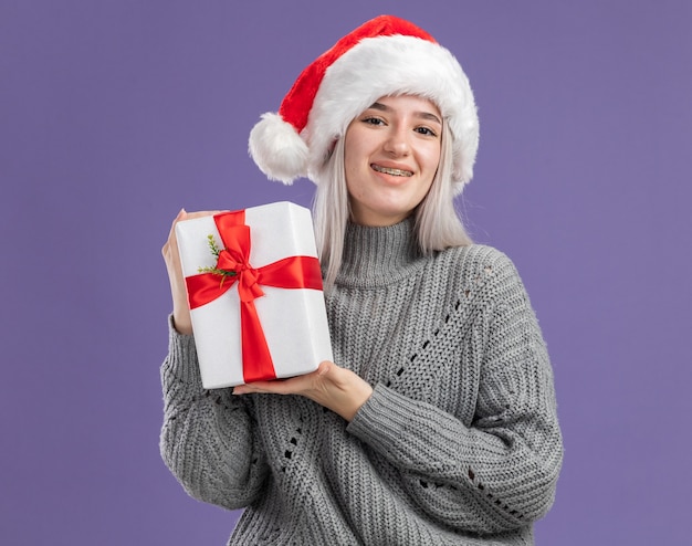 Junge blonde Frau in Winterpullover und Weihnachtsmütze, die ein Geschenk mit einem Lächeln auf einem glücklichen Gesicht hält, das über lila Wand steht