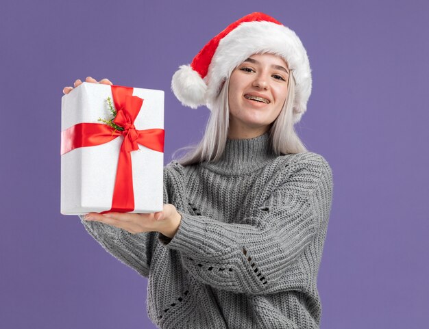 Junge blonde Frau in Winterpullover und Weihnachtsmütze, die ein Geschenk mit einem Lächeln auf einem glücklichen Gesicht hält, das über lila Wand steht